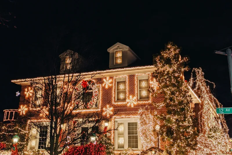 Two-story brick home at night with warm white Christmas lights outlining the roof, windows and a tall evergreen